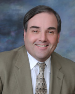 Chair member, James B. Turnbull. He is wearing a tan blazer with a white shirt and a tan tie.