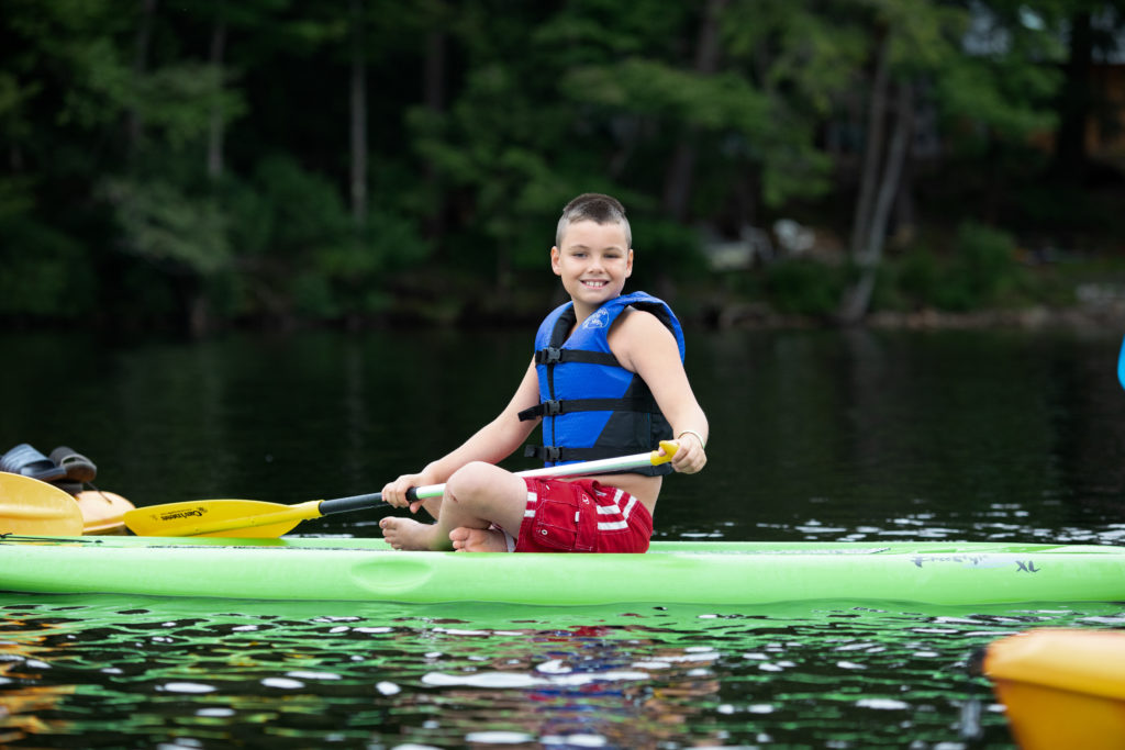 A visually impaired boy is sitting on a green paddle board in the middle of a lake
