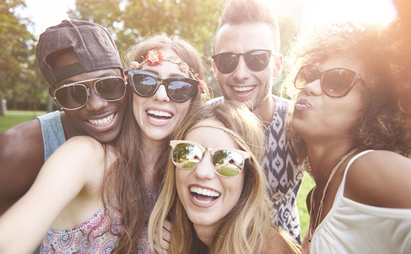 A group of people wearing sunglasses smiling. A sun flare is to the top right of them